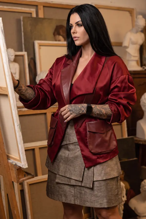 Woman in an oxblood satin blazer with leather lapels and a tiered tweed mini skirt beside an easel in a studio