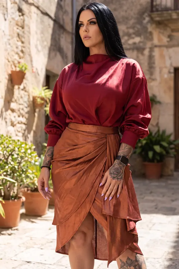 Woman in an oxblood satin blouse with blouson sleeves and a copper satin wrap skirt standing in a sunlit stone courtyard