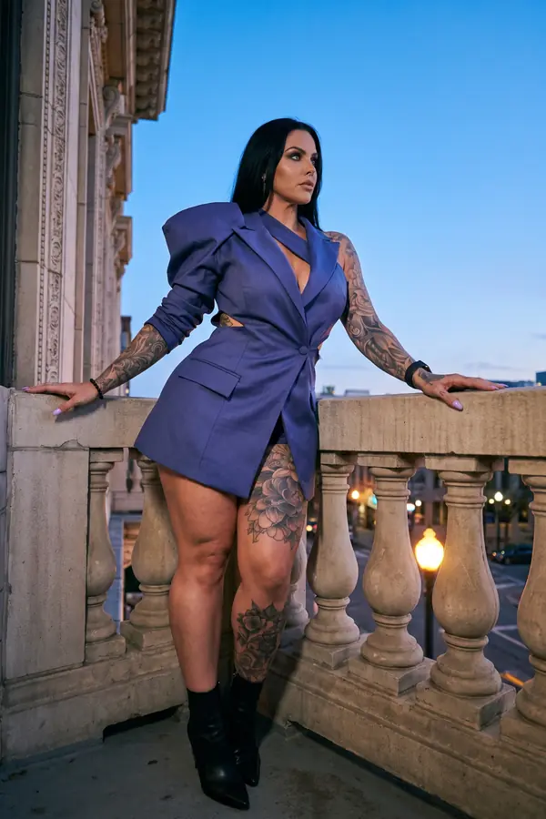 Woman in a violet-blue blazer dress with one puff sleeve and black ankle boots leaning on a stone balcony at dusk