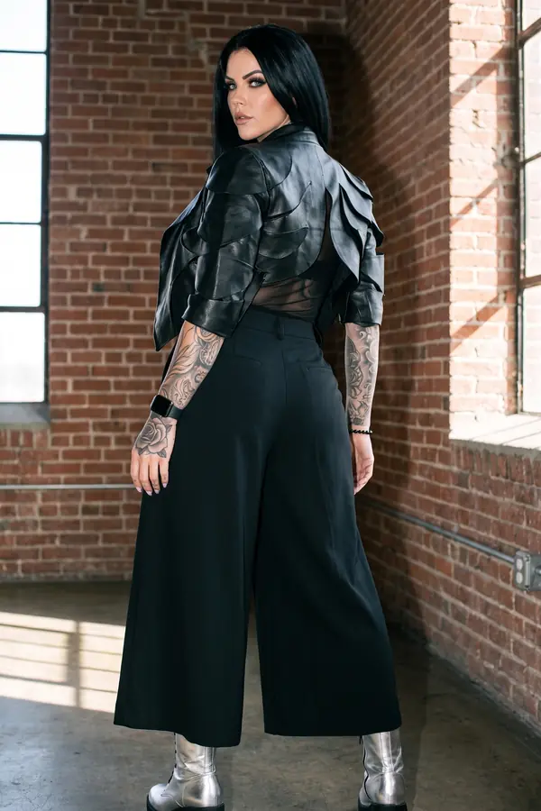 Woman seen from behind in a black petal-panel leather jacket, mesh top, wide-leg culottes and silver boots in a brick loft