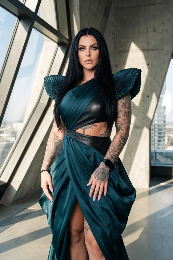 Woman in a petrol satin and black leather draped gown with pleated fan shoulders beside tall angled windows