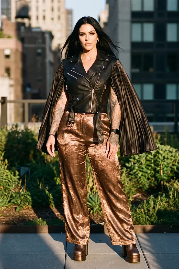 Woman in a cropped black leather biker vest with pleated cape sleeves and bronze metallic wide-leg trousers on a rooftop