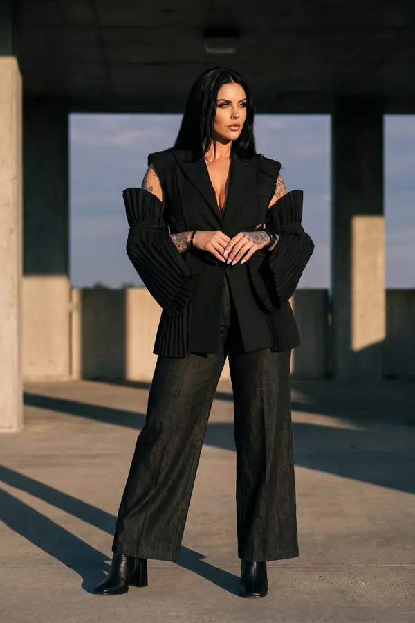 Woman in a sleeveless black blazer with pleated bell sleeves and black denim wide-leg trousers in a car park