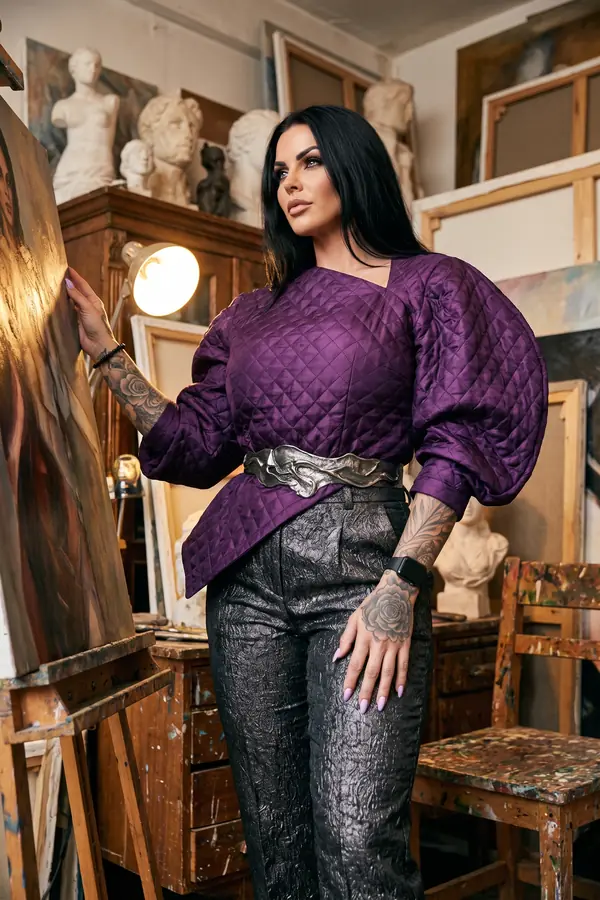 Woman in a purple quilted top with balloon sleeves, a sculpted silver belt and black metallic trousers in an artist's studio