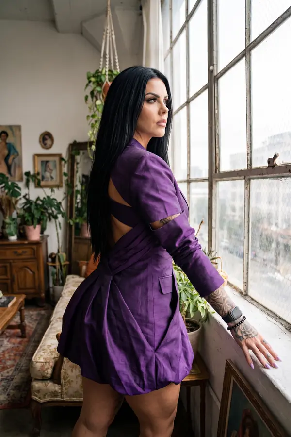 Woman seen from behind in a violet silk blazer dress with crossed back straps and a bubble hem, at a loft window