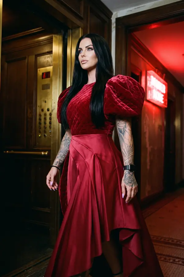Woman standing by a brass hotel elevator in a crimson quilted velvet puff-sleeve top and a red satin high-low skirt