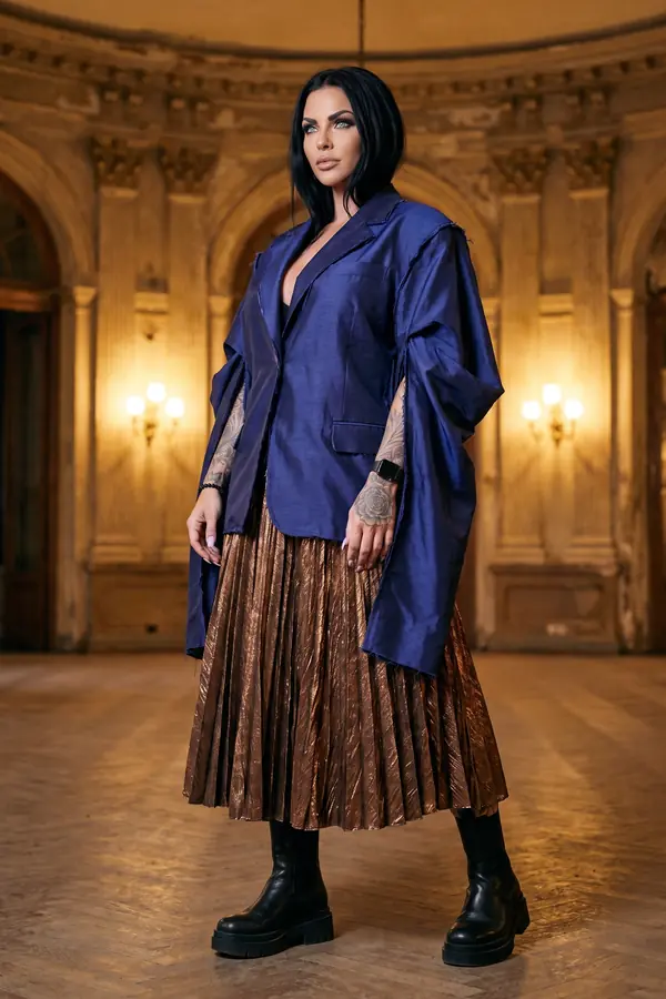 Woman in an oversized indigo silk blazer with raw edges and a bronze pleated midi skirt in a grand hall