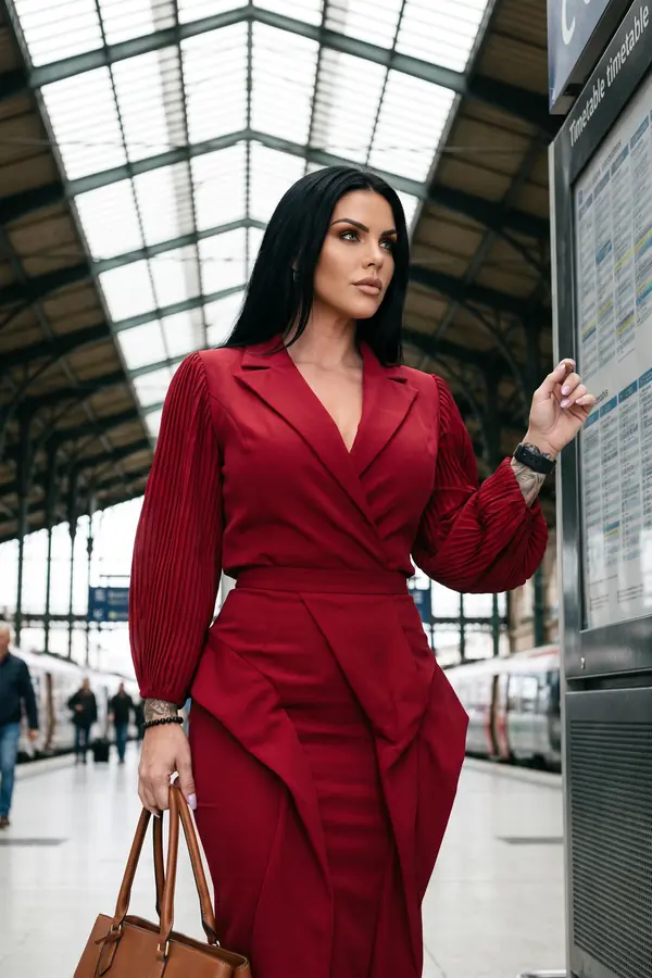 Woman in a red crepe wrap dress with pleated bishop sleeves holding a tan leather tote on a station platform
