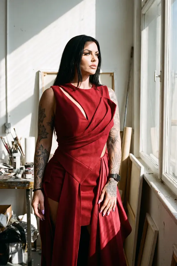 Woman in a sleeveless red draped dress with long panels and a slash cut-out by a sunny loft window