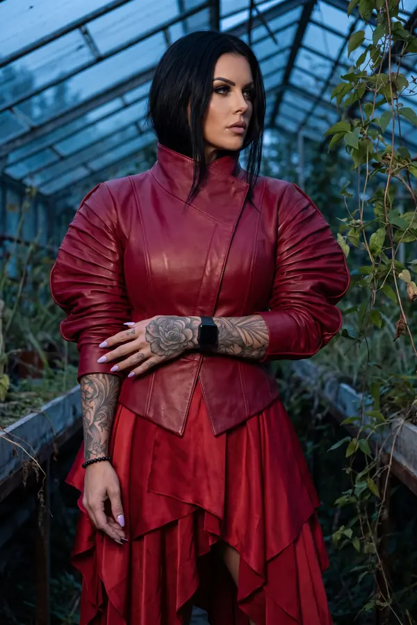 Woman in a crimson leather jacket with ruched balloon sleeves and a tiered red satin skirt standing in a greenhouse at dusk