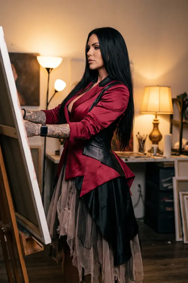 Woman in a crimson satin blazer with leather lapels and corset belt over a torn tulle skirt, painting at an easel