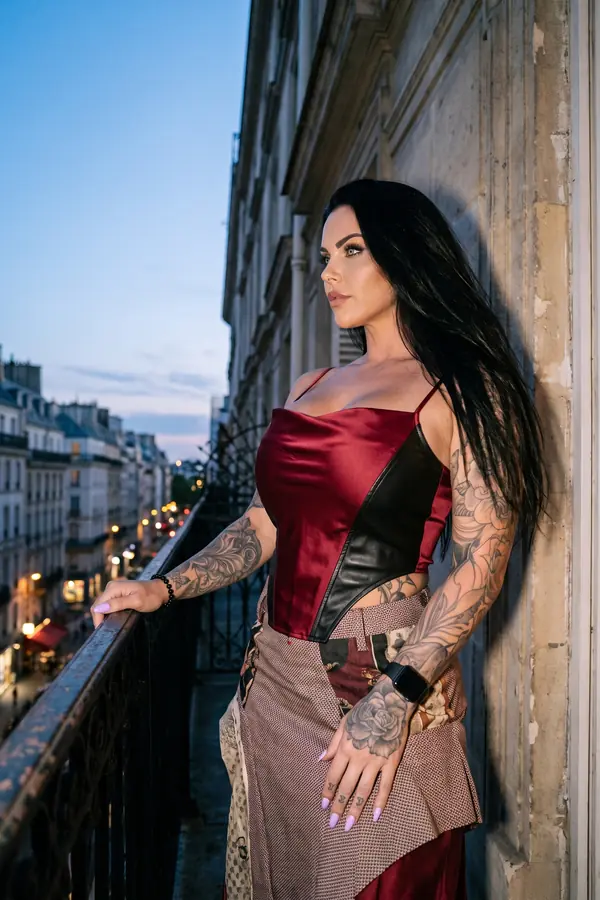 Woman in a red satin corset top and long patchwork wrap skirt leaning on a wrought-iron balcony at dusk