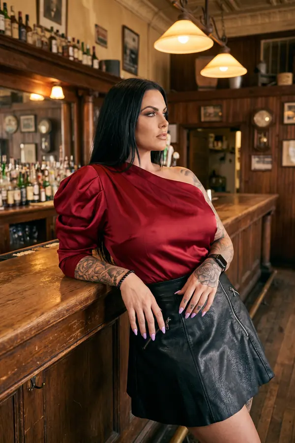 Woman in a red satin one-shoulder top with a puff sleeve and a black faux-leather zip mini skirt leaning on a wooden bar