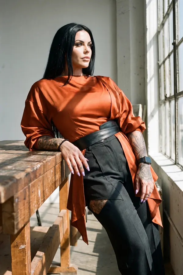 Woman with long black hair in a rust satin draped top and black cut-out trousers leaning on a workbench by a window