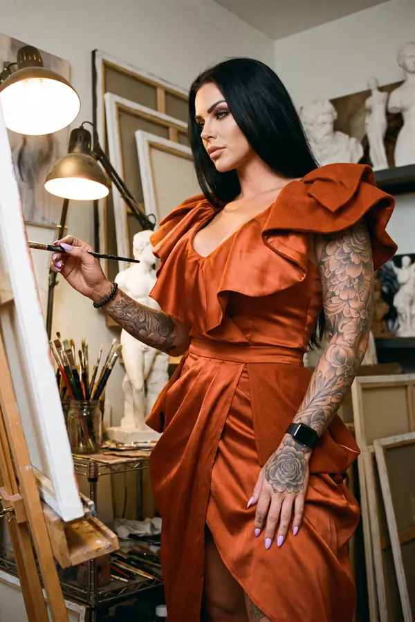 Woman in a rust satin ruffle dress with a draped wrap skirt painting at an easel in a lamp-lit artist's studio