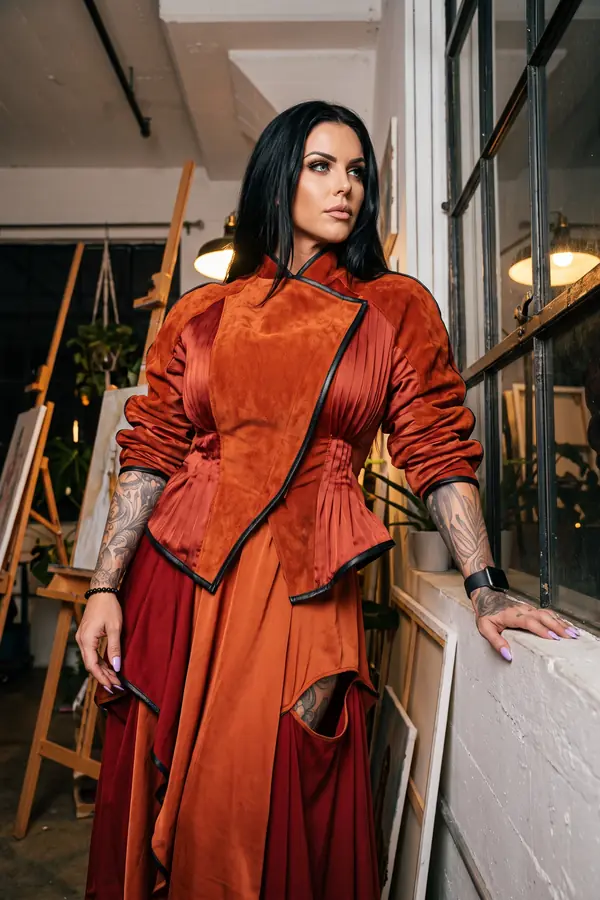 Woman in a rust suede jacket with pleated satin panels and a layered satin skirt leaning on a window sill in a loft studio