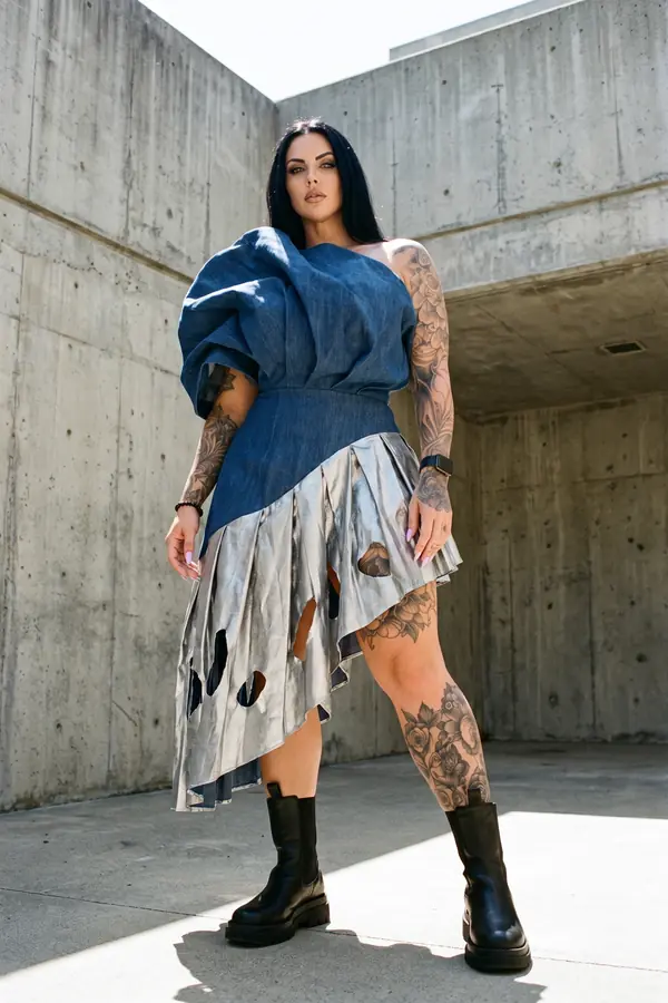 Woman in a sculpted denim one-shoulder dress with a silver pleated cut-out skirt and chunky boots in a concrete courtyard