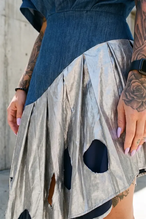 Woman in a sculpted denim one-shoulder dress with a silver pleated cut-out skirt and chunky boots in a concrete courtyard