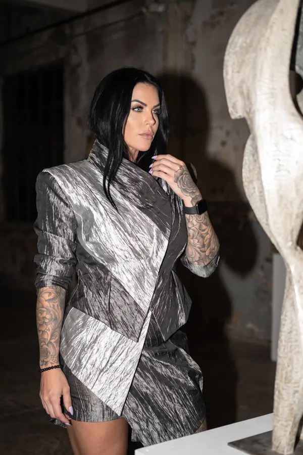 Woman in a silver crinkled metallic wrap mini dress with a funnel collar beside a pale stone sculpture in a derelict hall