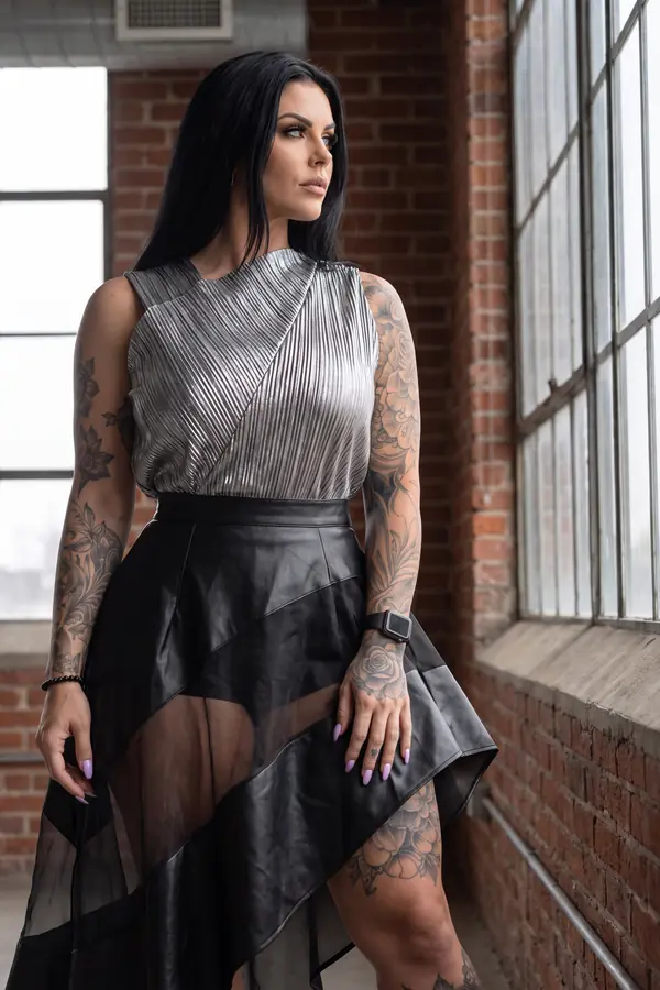 Woman in a silver pleated metallic top and black leather and organza high-low skirt by a steel window in a brick loft