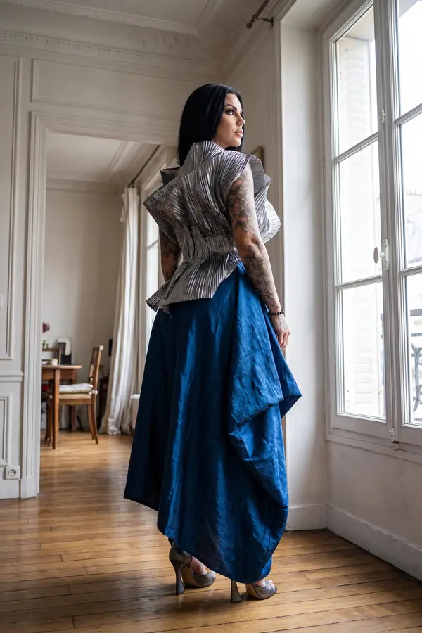 Woman seen from behind in a silver pleated metallic top and draped cobalt taffeta skirt by a tall window in an apartment