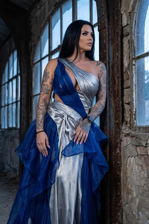Woman in a silver satin one-shoulder gown with cobalt organza ruffles standing by a rusted iron column in a derelict hall