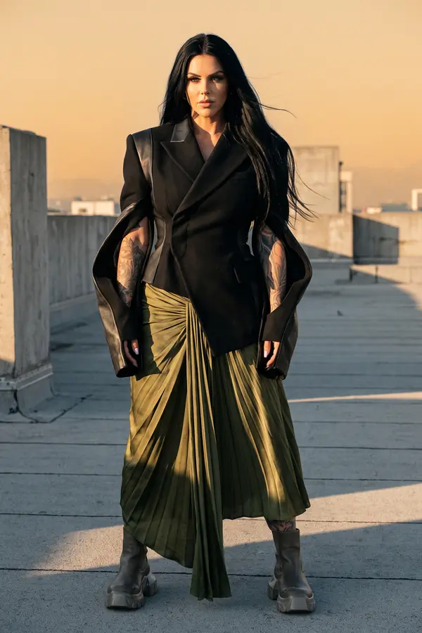 Woman in a black wool blazer with open split leather-lined sleeves and an olive pleated satin skirt on a rooftop at sunset