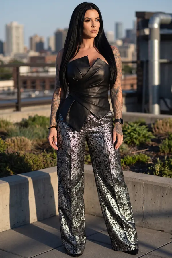 Woman in a strapless black leather bustier and wide silver brocade trousers on a rooftop garden at golden hour