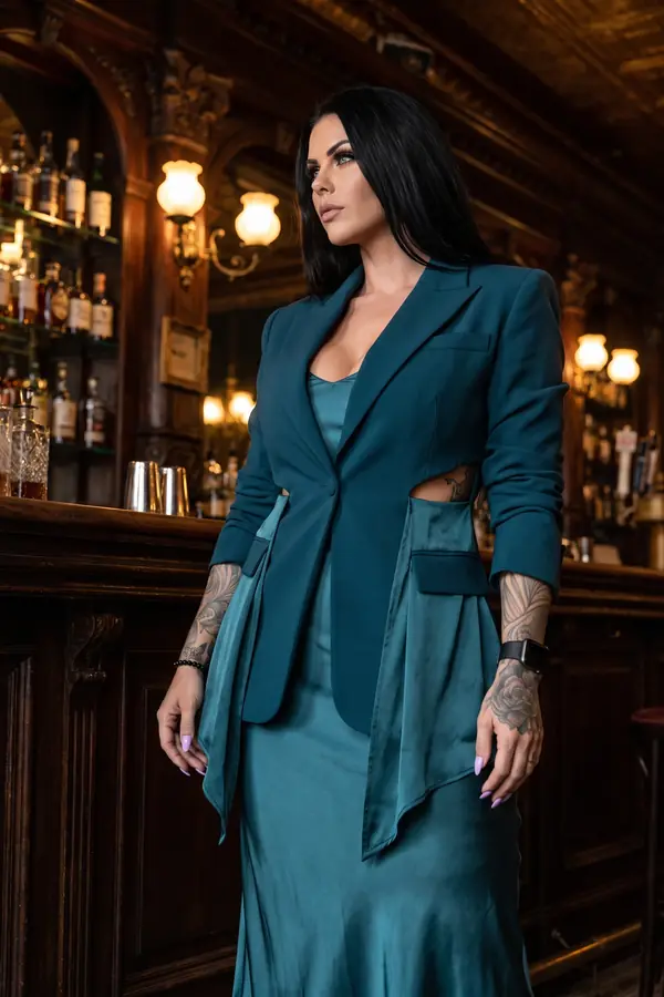 Woman in a petrol blazer with waist cut-outs over a teal satin slip dress standing at a wood-panelled whisky bar