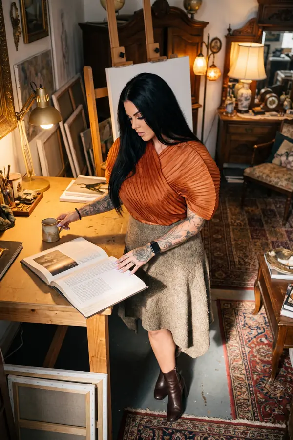 Woman reading at a desk in a terracotta pleated fan-sleeve top and taupe wool skirt in a room of lamps and antiques