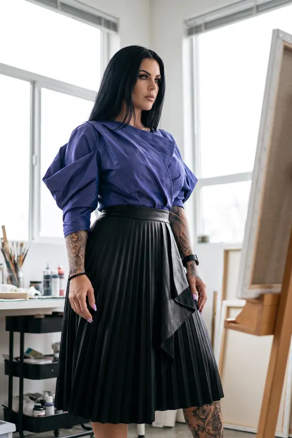 Woman in a violet taffeta draped top with puff sleeves and a black pleated faux leather midi skirt in a painting studio