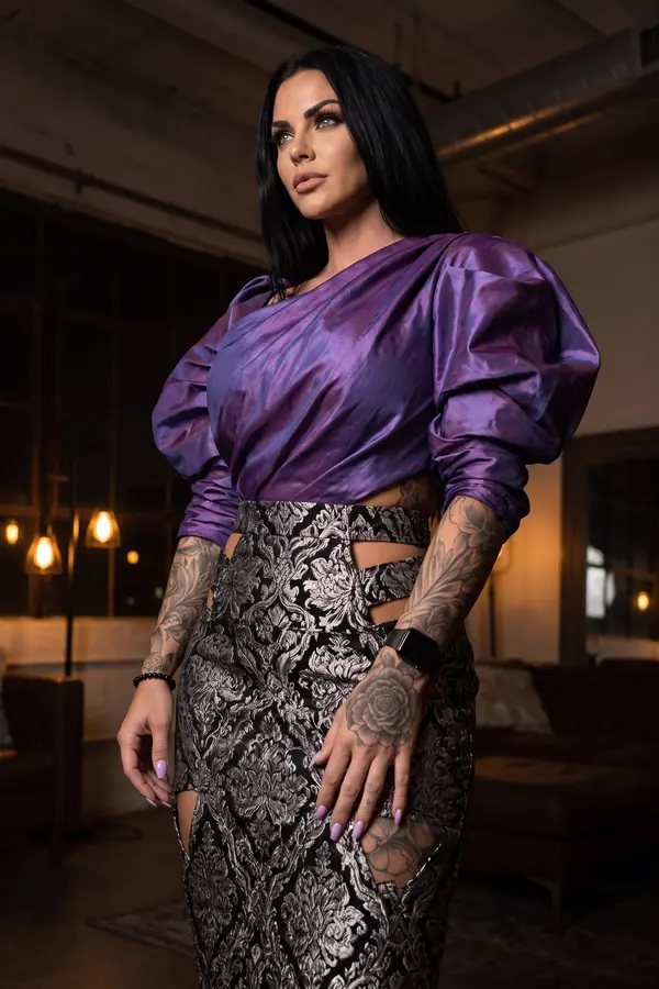 Woman in a violet taffeta one-shoulder top with balloon sleeves and a black and silver damask pencil skirt in a lounge